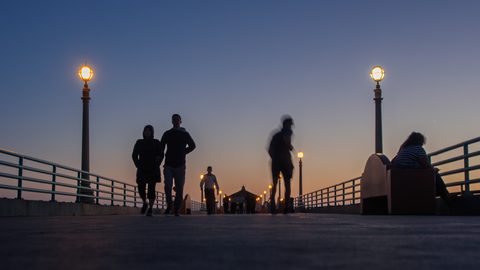 Silhouetted figures walking along a pier lit by lamp posts at dusk.