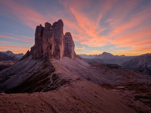 The jagged peaks of Tre Cime di Lavaredo lit orange at sunset beneath a sky streaked with pink clouds.