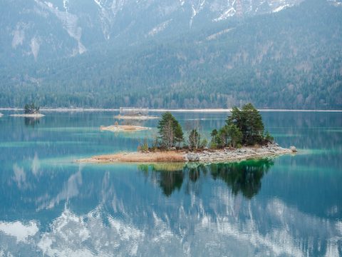 A small tree-covered island in a calm turquoise lake, mirrored in the still water with mountains beyond.