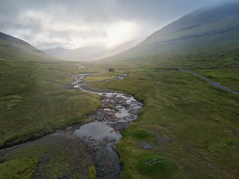 Aerial view of a winding river cutting through a green Faroese valley, with a lone house beside the water and mist rolling over the mountains beyond.