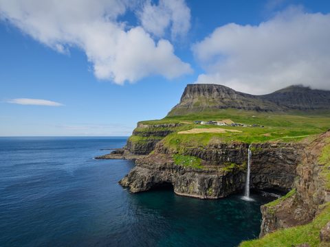 A waterfall dropping from green sea cliffs directly into the ocean, with a small village on the headland above.