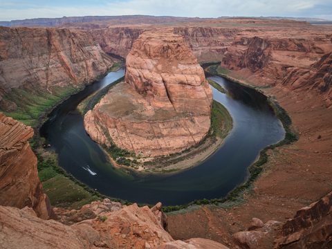 A dramatic horseshoe-shaped bend of the Colorado River carving through red sandstone canyon walls, with a small boat on the water.