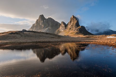 The jagged peak of Eystrahorn mountain reflected in a still pool, lit by warm evening light with clouds drifting past the summit.
