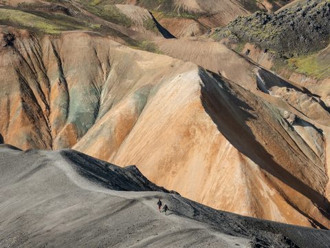 Two hikers walking a narrow ridge trail between striped rhyolite mountains in Landmannalaugar.