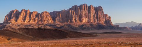 Panoramic view of the striated red sandstone cliffs of a jebel rising above the desert floor of Wadi Rum, lit gold by low sunlight.