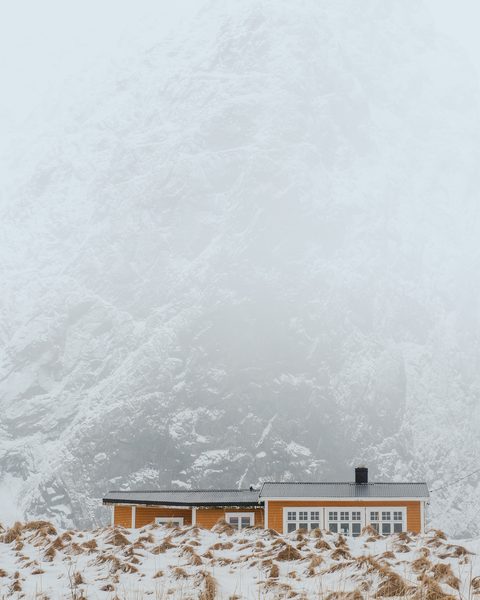 A small orange cabin at the base of a snow-covered mountain shrouded in fog.