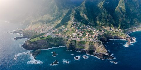 An aerial view of a coastal village perched on cliffs between green mountains and the sea.