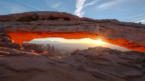 The sun bursts through the base of Mesa Arch at sunrise, lighting the underside of the arch orange against distant red-rock mesas.