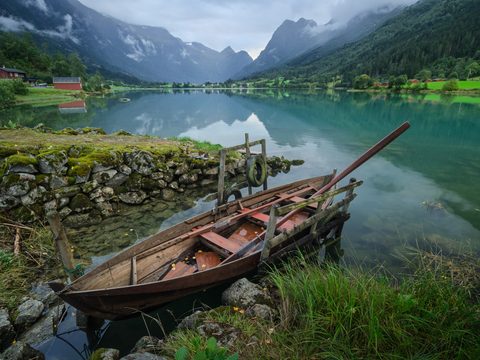 A weathered wooden rowboat tied up on a mossy stone shore, with a still turquoise lake and steep mountains behind it.