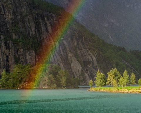 A vivid rainbow arcing down a steep cliff face into a turquoise fjord.