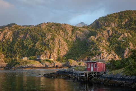A small red fishing hut on a wooden dock at the edge of a fjord, backed by rocky, tree-covered hills in evening light.