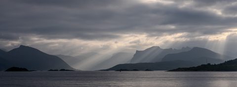 Shafts of sunlight breaking through heavy clouds over the silhouetted mountains and islands of Senja.