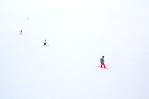 Skiers descending a wide, empty white ski slope, seen from above.